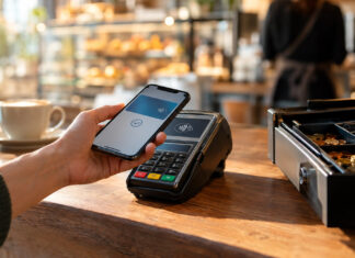 Hand hält Smartphone an ein Kartenterminal an einer Café-Kasse im warmen Tageslicht.