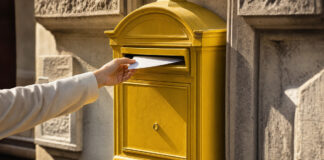 Eine Hand wirft einen einzelnen Brief in einen gelben Briefkasten an einer sonnigen Altbaufassade