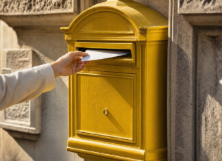 Eine Hand wirft einen einzelnen Brief in einen gelben Briefkasten an einer sonnigen Altbaufassade