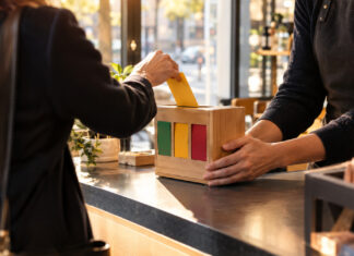 Hand am Tresen mit Feedback-Kasten, eine Person wirft von hinten eine Karte ein