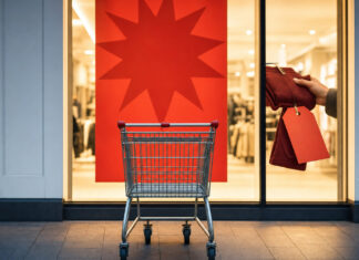 Einkaufswagen vor Schaufenster mit Sale-Plakat und Hand am reduzierten Produkt