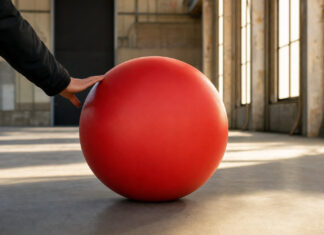 Ein roter Gummiball liegt in einer Werkhalle und wird von hinten von einer Hand angestoßen.