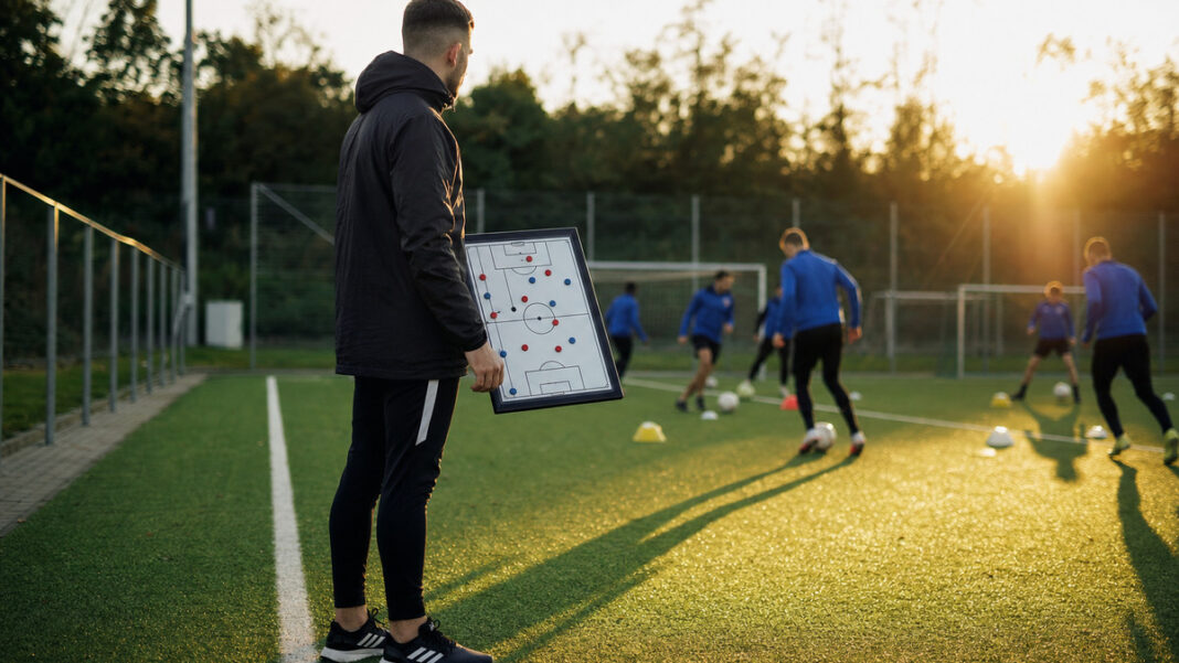 Trainerassistent am Fußballplatz Trainerassistent mit Taktikboard am Fußballplatz, im Hintergrund trainiert eine kleine Gruppe bei Abendlicht.