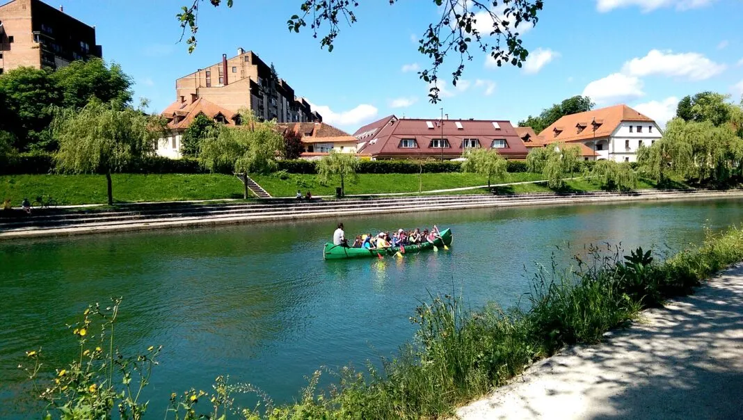 Grünes Kanu auf der Ljubljanica, dahinter Uferstufen, Trauerweiden und Wohnhäuser