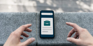 Smartphone mit reduzierter Jobsuche auf Steinmauer, Hände greifen danach im Tageslicht