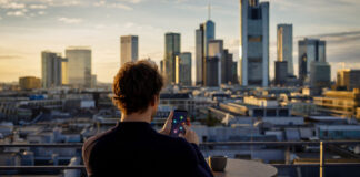 Person verbindet auf einer Dachterrasse farbige Ortsmarken per Faden, Stadt im Abendlicht