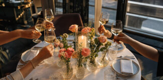 Runder Dinner-Tisch mit Kerzen und Blumen, darum herum anstoßende Frauenhände im warmen Licht.