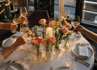 Runder Dinner-Tisch mit Kerzen und Blumen, darum herum anstoßende Frauenhände im warmen Licht.