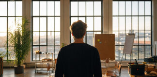 Junger Gründer von hinten vor einer Fensterfront im Betahaus, mit kleinem Workshop-Bereich und Morgenlicht.