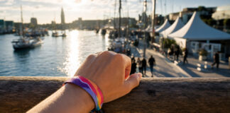 Hand spannt ein Festivalarmband am Hafenkai, dahinter Zelte, Masten und Wasser im Sommerlicht.