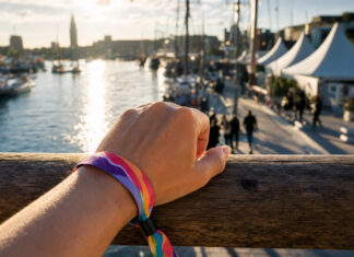 5 Gründe das Waterkant Festival in Kiel zu besuchen Hand spannt ein Festivalarmband am Hafenkai, dahinter Zelte, Masten und Wasser im Sommerlicht.