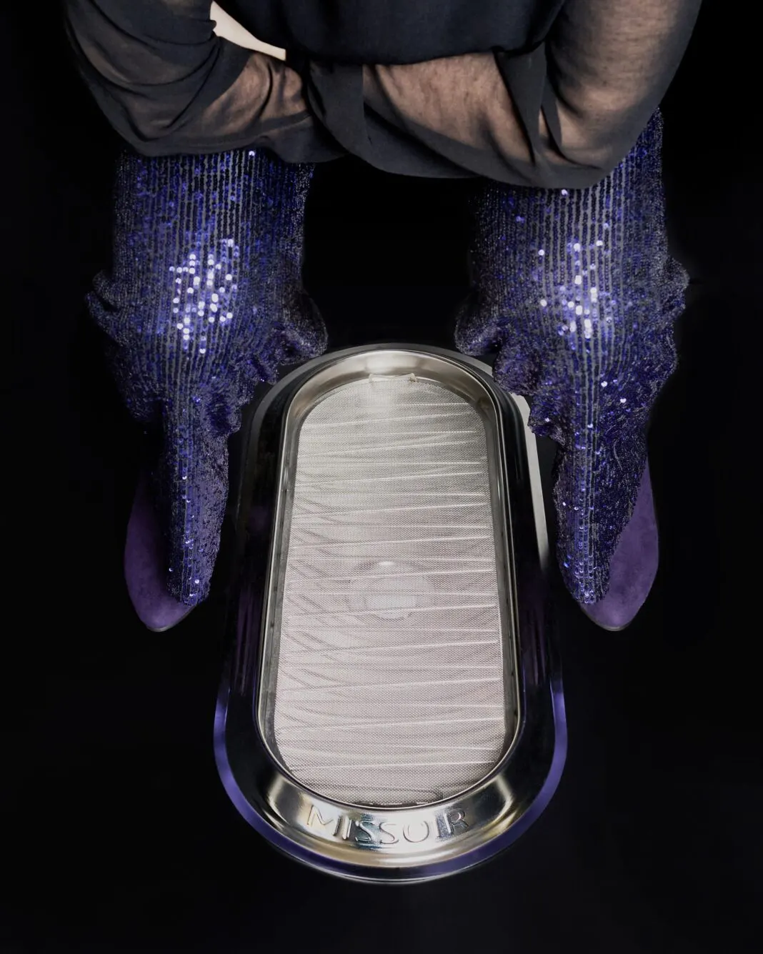 Blick von oben auf ein Hockurinal aus Edelstahl, davor steht eine Person in Hose und Stiefeln in der Hocke