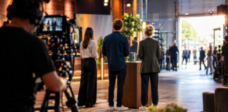 Gründerteam mit Produktkarton in einer TV-Studio-Halle, Kamerastand und helles Licht