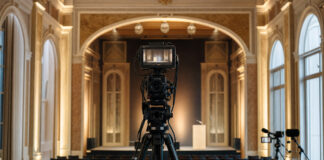 Kamera auf Stativ vor Bühne in Kongresssaal, daneben Livestream-Setup mit warmem Tageslicht