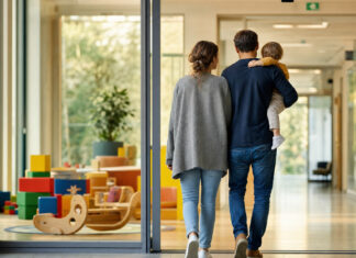 KW 12 | Corona-Krise: Krankenhäuser hoffen auf Hilfe in der Kinderbetreuung Eltern mit Kleinkind am Eingang eines Krankenhauses, im Hintergrund eine leere Kinderbetreuungsecke.
