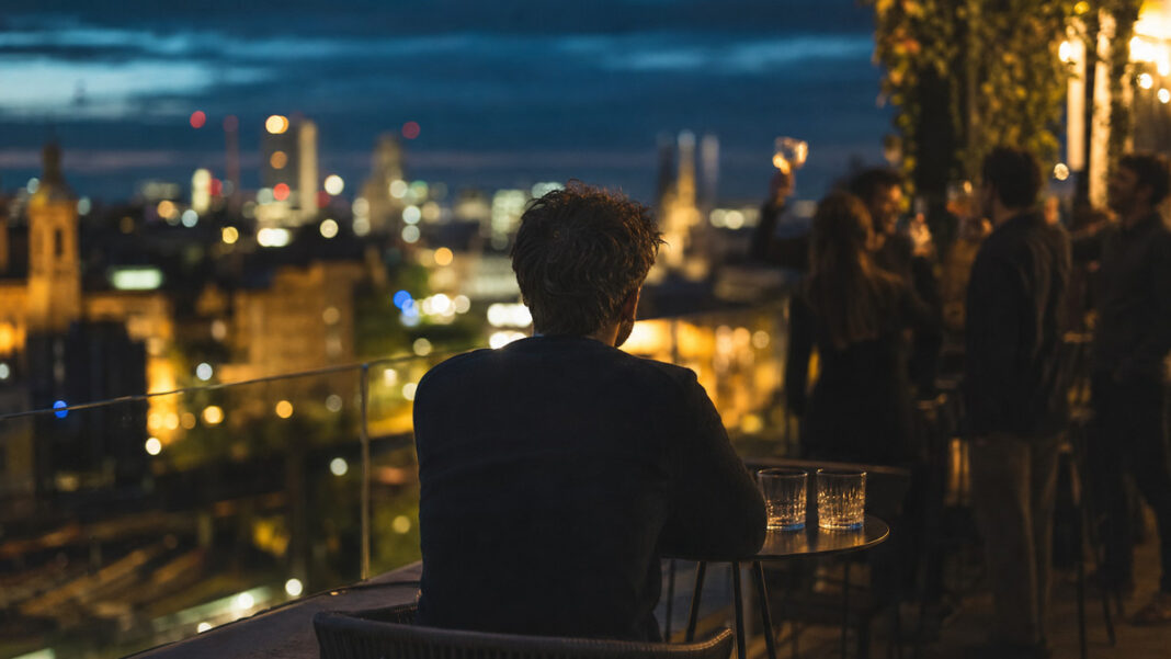 Gründer sitzt allein auf Dachterrasse, im Hintergrund lachen Freunde an einer Bar