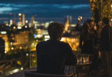 Mit Freunden gründen oder saufen? Gründer sitzt allein auf Dachterrasse, im Hintergrund lachen Freunde an einer Bar