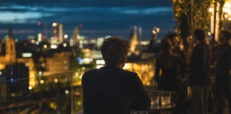 Gründer sitzt allein auf Dachterrasse, im Hintergrund lachen Freunde an einer Bar