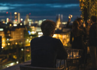 Gründer sitzt allein auf Dachterrasse, im Hintergrund lachen Freunde an einer Bar