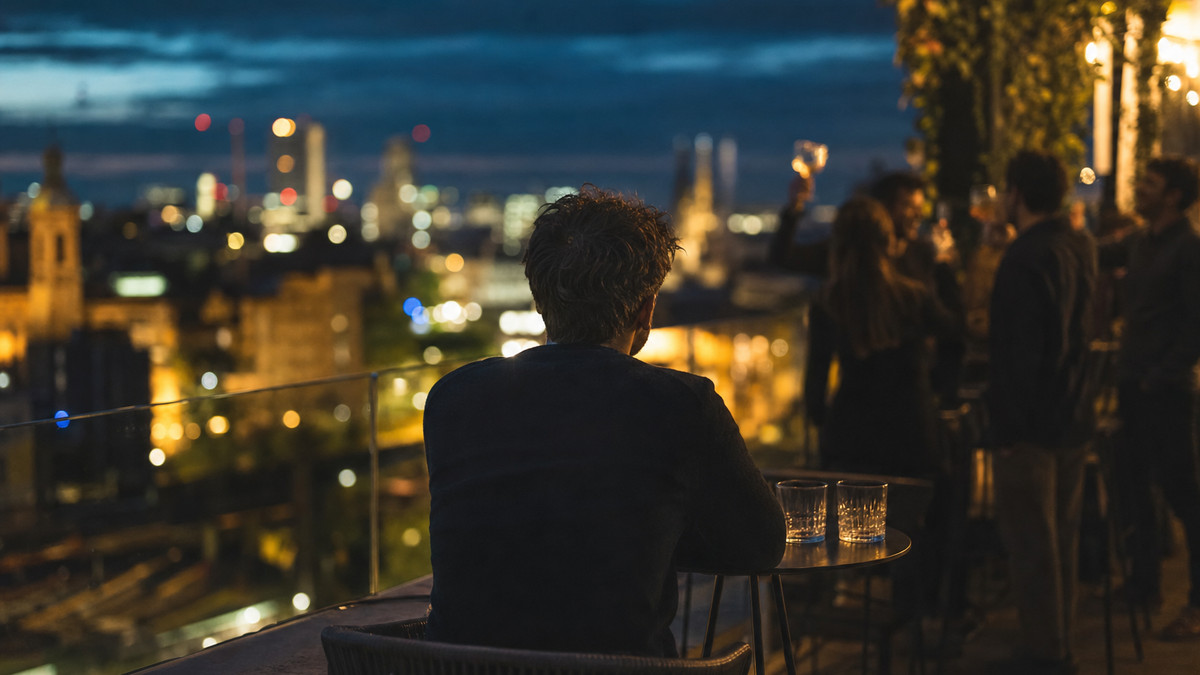 Mit Freunden gründen oder saufen? Gründer sitzt allein auf Dachterrasse, im Hintergrund lachen Freunde an einer Bar