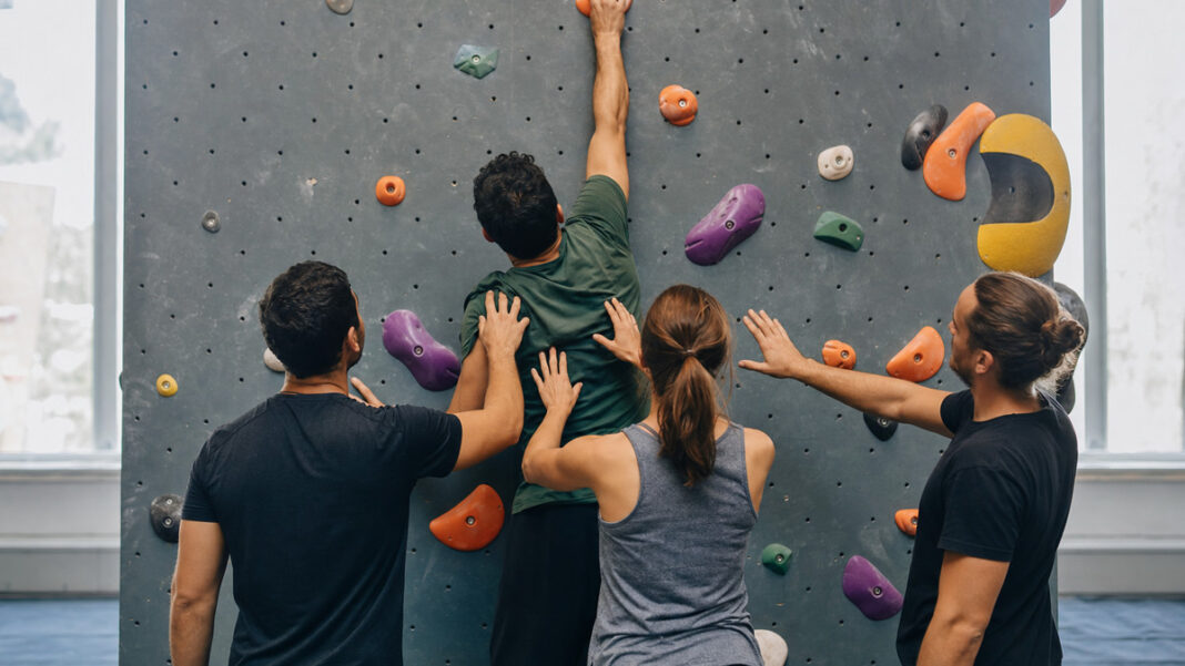 Vier Personen an einer Boulderwand, Hände an Griffen und ein letzter Griff oben im Licht.