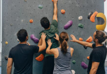 Vier Personen an einer Boulderwand, Hände an Griffen und ein letzter Griff oben im Licht.