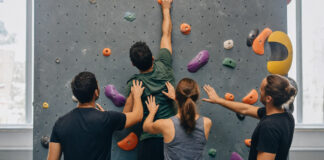 Vier Personen an einer Boulderwand, Hände an Griffen und ein letzter Griff oben im Licht.