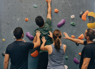 Vier Personen an einer Boulderwand, Hände an Griffen und ein letzter Griff oben im Licht.