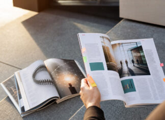 Auf einer Treppenstufe liegen mehrere Magazine, eine Hand hält ein weiteres Heft im Sonnenlicht.