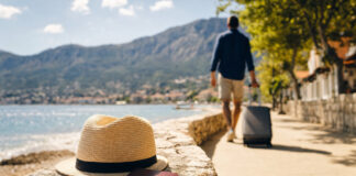 Person mit Rollkoffer an der Strandpromenade bei sonnigem Licht, Reisepass und Sonnenhut im Vordergrund.