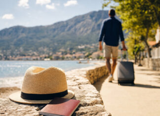 Person mit Rollkoffer an der Strandpromenade bei sonnigem Licht, Reisepass und Sonnenhut im Vordergrund.