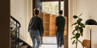 Zwei Gründer tragen eine Holzregalwand in einen hellen Altbau, daneben stehen Stuhl, Pflanze und Lampe.
