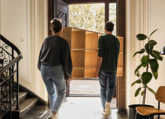 Zwei Gründer tragen eine Holzregalwand in einen hellen Altbau, daneben stehen Stuhl, Pflanze und Lampe.