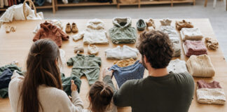 Sortierte Kinderkleidung auf einem Holztisch, eine Familie betrachtet die Stücke von hinten im Tageslicht.