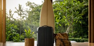Gepackter Koffer mit Surfbrett und tropische Pflanze auf einer Bali-Terrasse im Morgenlicht
