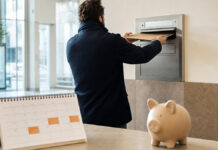 Gründer wirft eine Mappe in einen Wandbriefkasten, daneben Kalender und Sparschwein im Tageslicht.