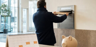 Gründer wirft eine Mappe in einen Wandbriefkasten, daneben Kalender und Sparschwein im Tageslicht.