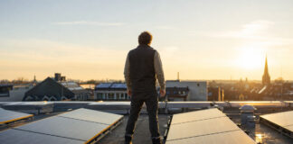 Gründer prüft auf dem Dach seiner Manufaktur eine neue Solaranlage, dahinter Stadtdächer im Morgenlicht