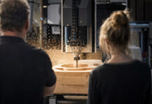 KI im Mittelstand verkaufen 2026: Das B2B-Playbook für Startups Werkstattmeister und Entwicklerin beobachten in einer Schreinerei eine CNC-Fräse bei der Arbeit