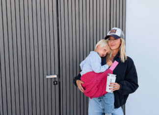 Eine Frau mit Basecap trägt ein Kleinkind in einer pinken monobag-Tasche vor einer grauen Holzwand