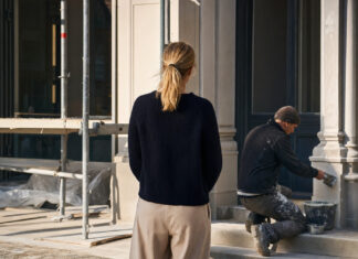 Gründerin vor einer Baustellenfassade, daneben ein Handwerker mit Kelle im warmen Tageslicht.