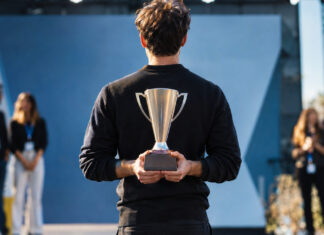 Gründer hält auf einer Startup-Bühne einen Siegerpokal, andere Teams stehen unscharf im Hintergrund.