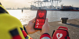 Hände halten ein Smartphone mit Notfall-App neben einem AED-Koffer am Hamburger Hafen