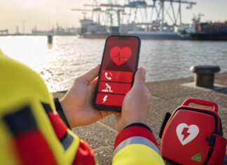 HAMBURG SCHOCKT – Eine Handy App, die Leben rettet. Erste Hilfe für Jedermann! Hände halten ein Smartphone mit Notfall-App neben einem AED-Koffer am Hamburger Hafen