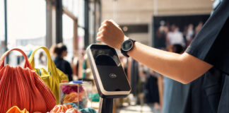 Kontaktloses Zahlungsterminal neben Mode-Accessoires, eine Hand hält eine Armbanduhr im warmen Tageslicht.