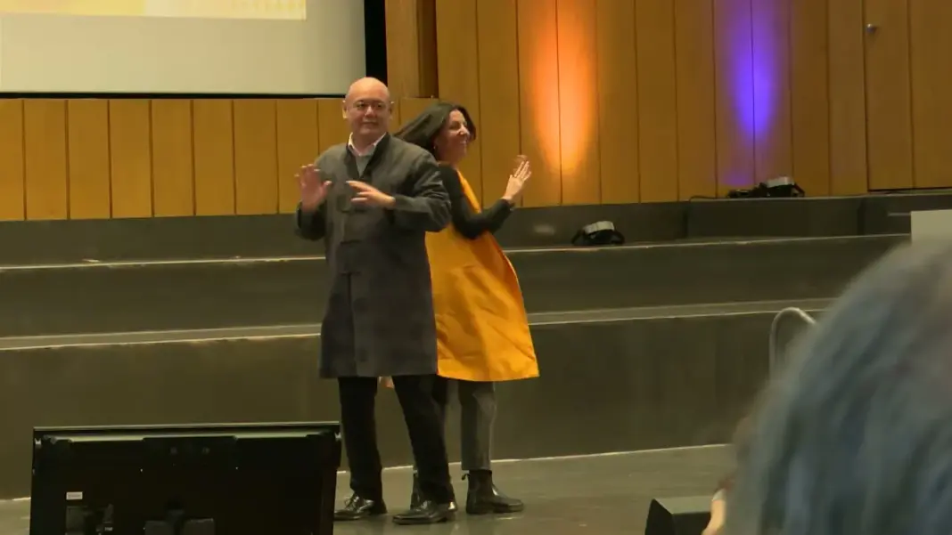 Awai Cheung und Moderatorin Dörte Roloff in gelber Weste machen auf der Bühne des Audimax eine Bewegungsübung vor