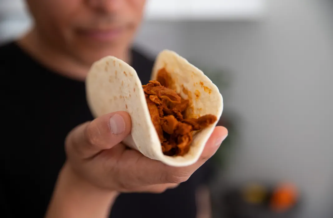 Eine Hand hält einen gefalteten Maistortilla-Taco mit rotbrauner Füllung in die Kamera