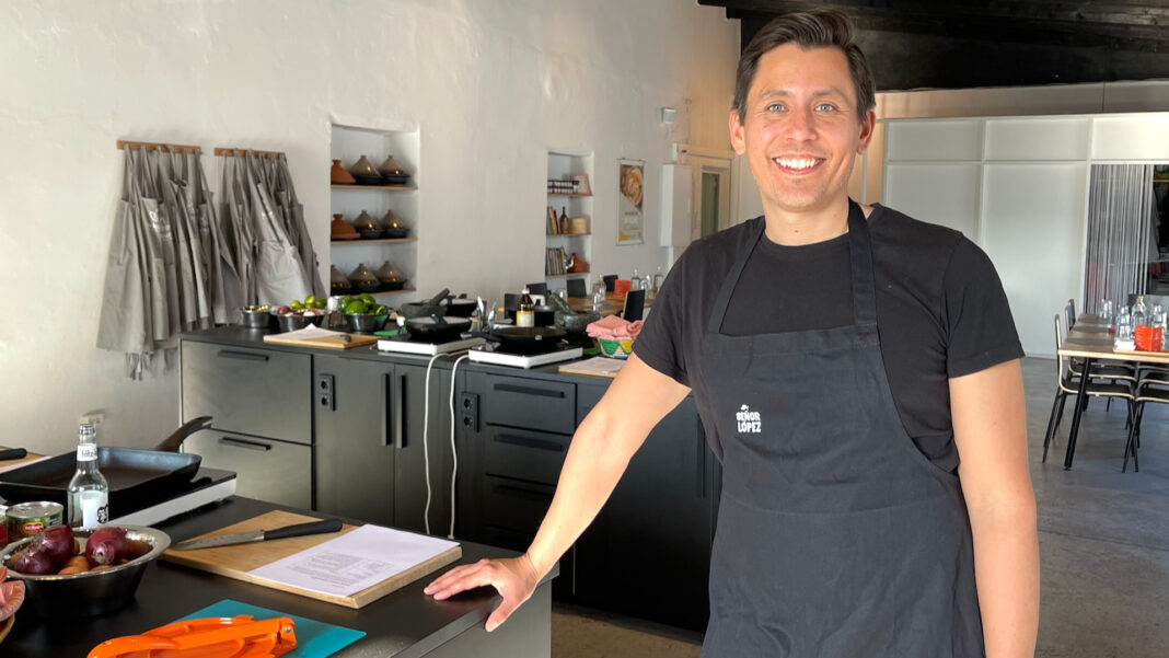 Mathyas López steht in Schürze mit Señor-López-Logo in einer Kochschulküche und lächelt in die Kamera