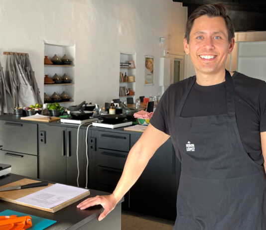 Mathyas López steht in Schürze mit Señor-López-Logo in einer Kochschulküche und lächelt in die Kamera