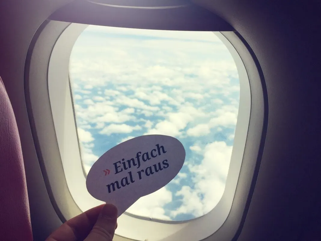 Hand hält eine Karte mit der Aufschrift Einfach mal raus vor ein Flugzeugfenster über den Wolken
